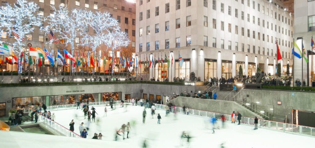 People ice skating at Rockefeller Center in New York City. The rink is surrounded by brightly lit trees and numerous international flags. The background features a large building with illuminated windows.