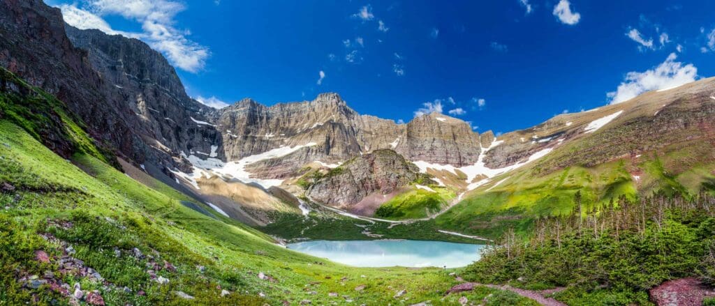 A panoramic view of a mountain landscape with a clear blue sky. Snow-dusted peaks rise above a lush green valley, surrounding a turquoise lake. Patches of snow are visible on the mountainside, with dense green forests in the foreground.