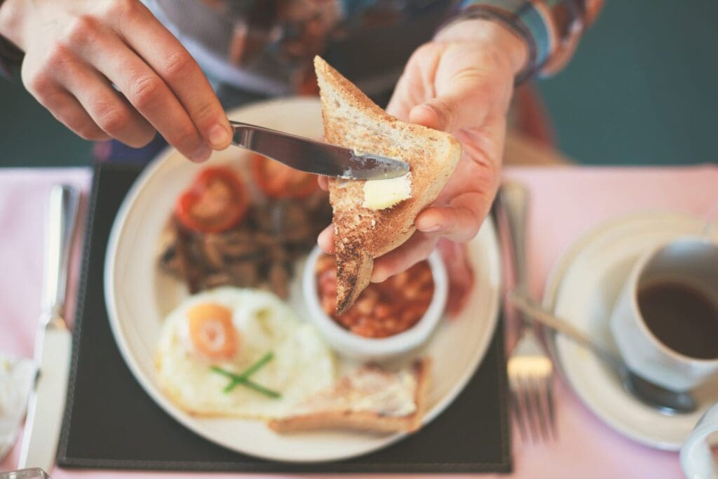 Person spreading butter on toast with a knife. Plate with fried egg, mushrooms, and baked beans in the background. Cup of coffee on the side. Silverware on the table. Cozy breakfast setting.