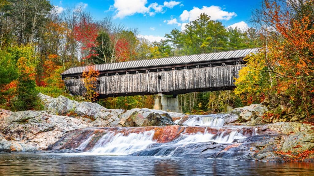 A rustic wooden covered bridge spans over a cascading waterfall surrounded by vibrant autumn foliage. The sky is blue with scattered clouds, and rocky terrain is visible in the foreground.
