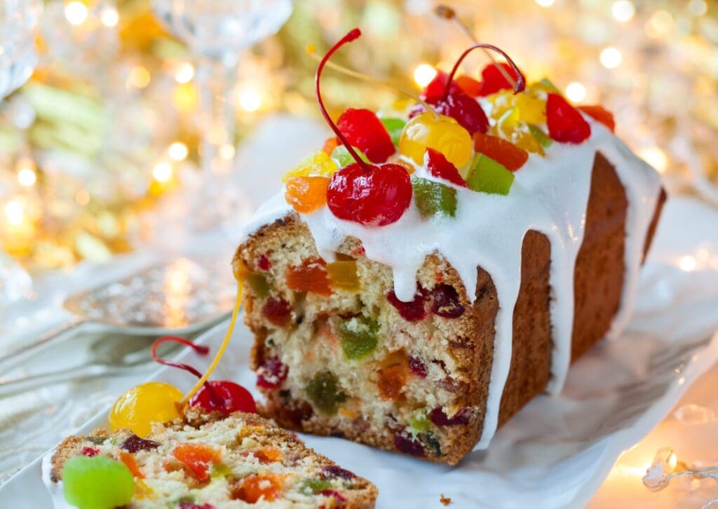 A festive fruitcake is topped with a white glaze and adorned with colorful candied fruits and cherries. The cake is sliced, revealing a rich, fruit-studded interior, set against a background of soft, warm holiday lights.