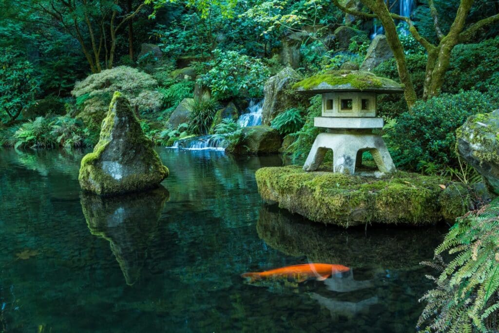 A tranquil Japanese garden features a koi pond with a bright orange fish swimming. A moss-covered stone lantern sits on a small island, surrounded by lush greenery and ferns. A gentle waterfall cascades in the background.