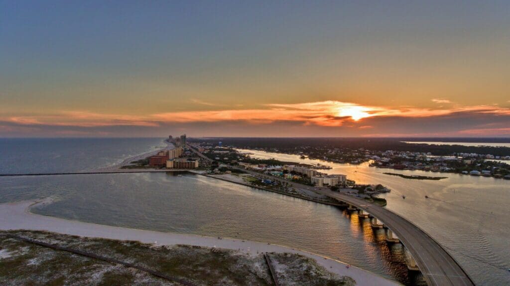Aerial view of a coastal city at sunset, featuring a bridge over water. The sun sets on the horizon with glowing orange and pink hues. Buildings line the shoreline on the left, while water and land are visible in the foreground.