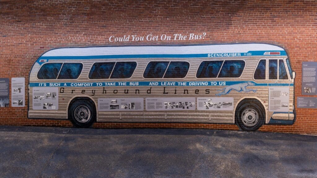A mural of a vintage Greyhound bus is painted on a brick wall. It features the slogan, Its such a comfort to take the bus...and leave the driving to us. Historical photos and text are integrated into the design, with the question Could You Get On The Bus? above.