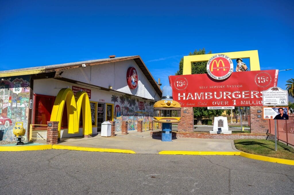Exterior of a retro-style fast-food restaurant with a large, classic McDonalds sign, displaying 15¢ HAMBURGERS and OVER 1 MILLION SOLD. The setting includes colorful murals and a hamburger-shaped statue.
