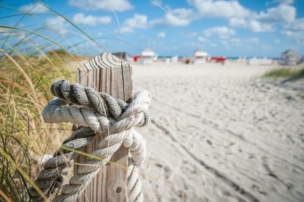 A wooden post wrapped with rope stands on a sandy beach with tall grasses. In the blurred background, red and white beach huts are visible under a partly cloudy blue sky.