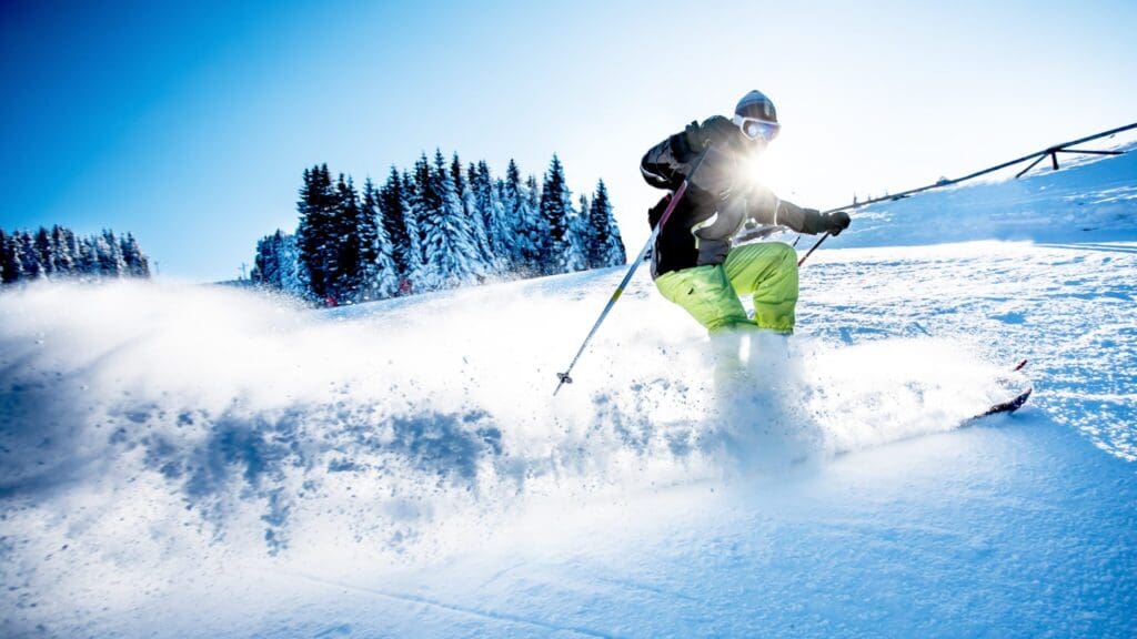 A skier in a grey jacket and bright green pants carves through fresh snow on a sunlit slope. Snow sprays in the air as they turn. Pine trees and a clear blue sky are visible in the background.