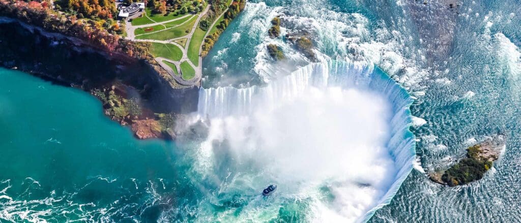 Aerial view of a large waterfall, with turquoise water cascading into a misty pool below. A small boat is near the base of the waterfall. Lush green landscape surrounds the top, with a park area visible.
