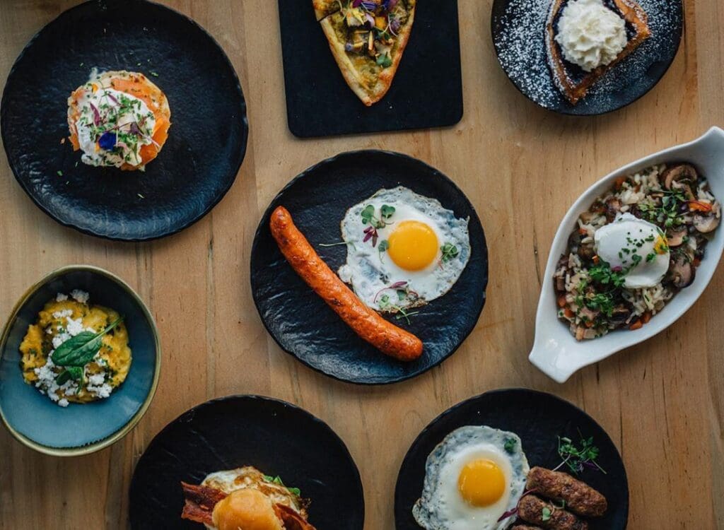 A wooden table set with various gourmet dishes on black plates, including a sausage with fried eggs, a slice of pizza, a dessert with whipped cream, and a dish with mushrooms and an egg. Garnishes and floral accents decorate some plates.