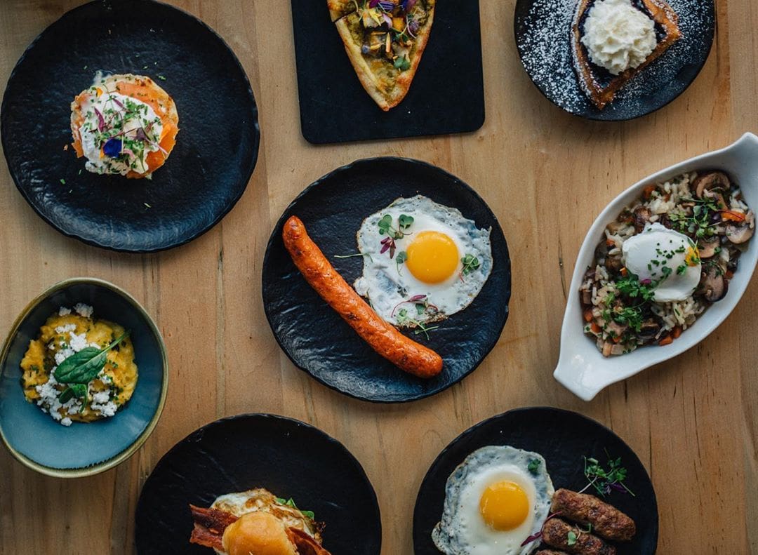 A wooden table set with various gourmet dishes on black plates, including a sausage with fried eggs, a slice of pizza, a dessert with whipped cream, and a dish with mushrooms and an egg. Garnishes and floral accents decorate some plates.