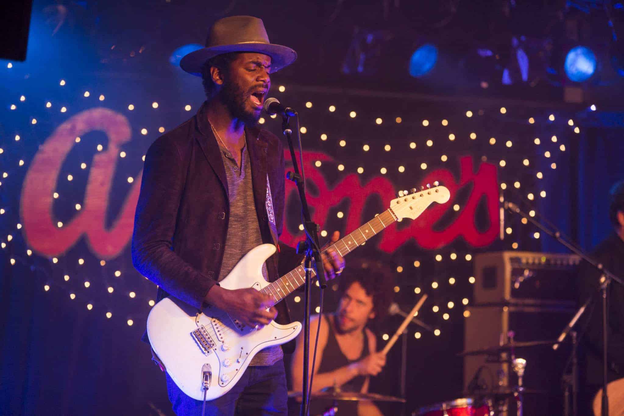 A musician wearing a hat plays an electric guitar and sings into a microphone on stage. A drummer is visible in the background. The stage is decorated with string lights and a sign reading Antones is partially visible.