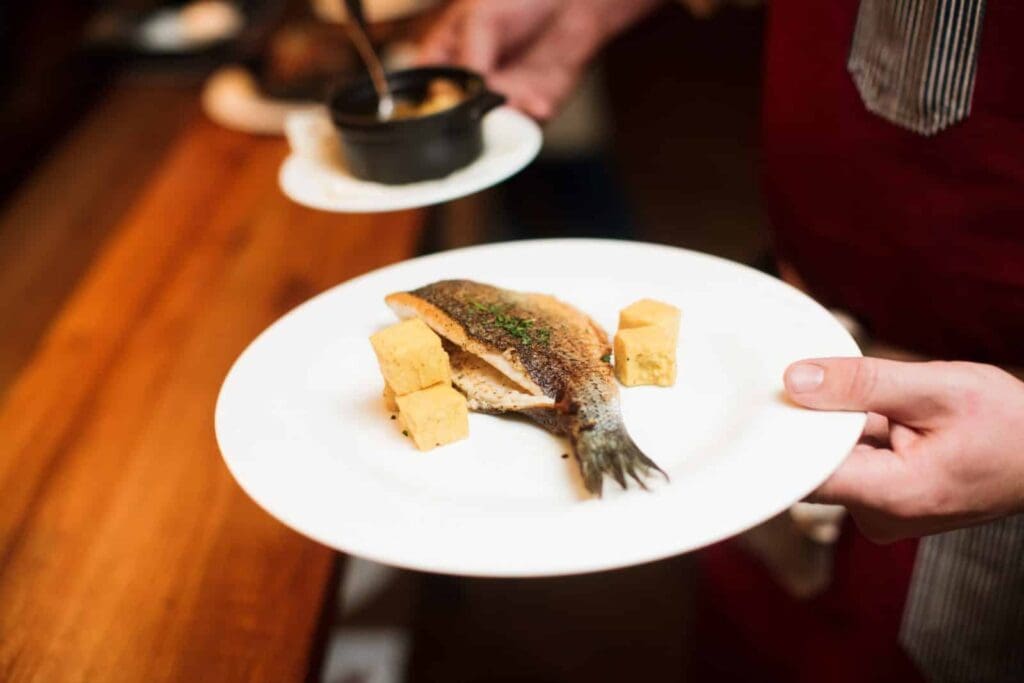 A person holding a white plate with a cooked fish garnished with herbs, accompanied by three golden-brown cubes, likely fried polenta. The person wears a red apron, and a small bowl is visible in the background.