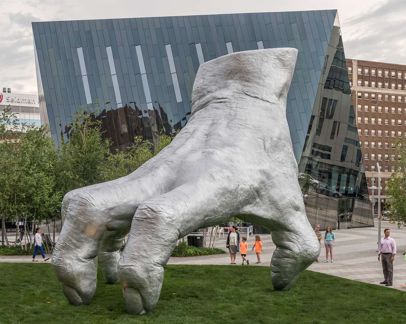 A large metallic sculpture of a foot with curled toes stands on a grassy area. In the background, there is a modern, angular glass building and several people walking nearby. Trees are scattered around the sculpture.
