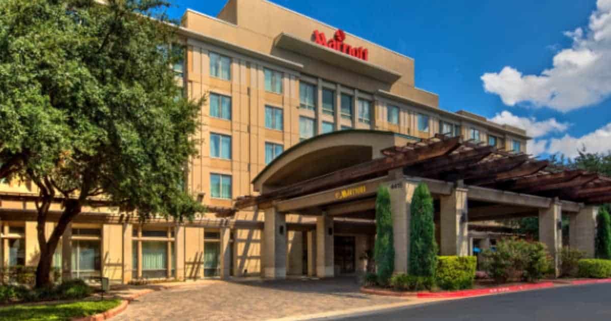 Exterior of a Marriott hotel with a beige facade and several floors. A covered entrance is surrounded by green trees and shrubs. The sky is partly cloudy, and a paved driveway leads to the main entrance.
