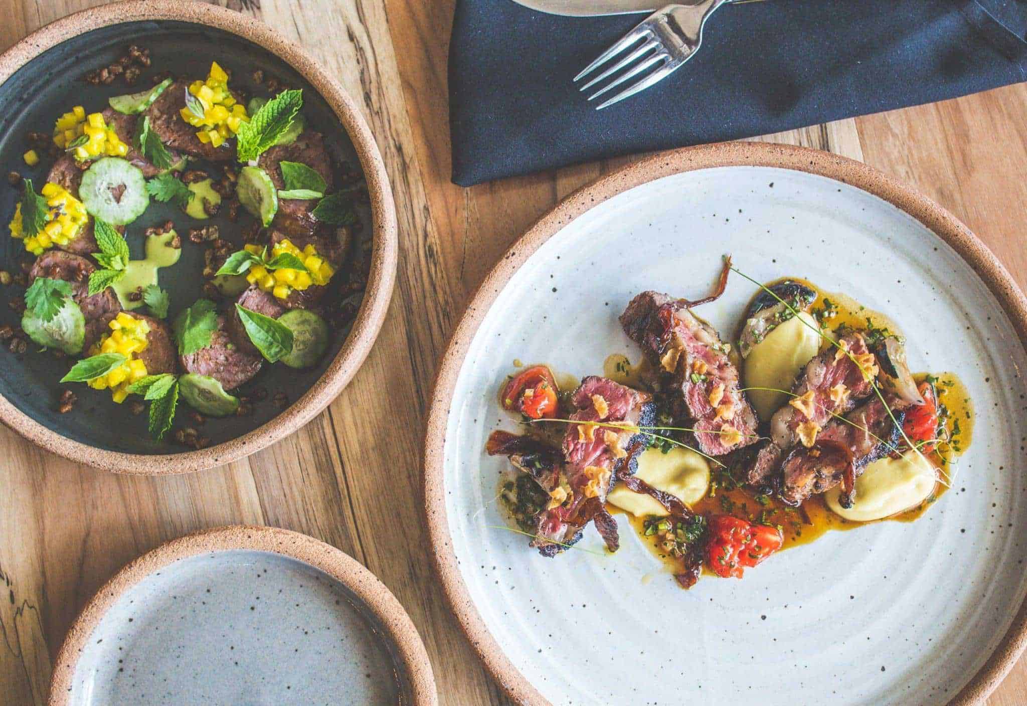 Two elegantly plated dishes on a wooden table. One plate has sliced meats garnished with yellow corn and herbs, the other features stuffed pasta with diced tomatoes and a drizzle of sauce. A fork and napkin are placed nearby.