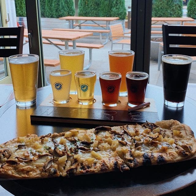 A table with a long flatbread topped with vegetables and cheese. Behind it, theres a wooden flight board holding five glasses of different colored beers. Outdoor seating with wooden benches and tables is visible through a window.
