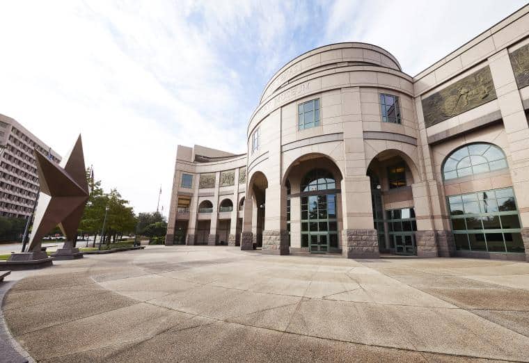 Wide-angle view of the Bob Bullock Texas State History Museum in Austin, Texas. The building features a curved facade with large arched windows and columns. A large bronze star sculpture stands to the left on the plaza. Sky is partly cloudy.
