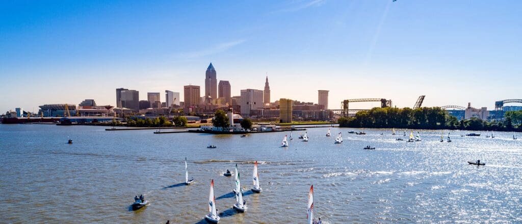Aerial view of a city skyline by the water with numerous sailboats on a sunny day. The horizon features tall buildings, and the foreground showcases a wide river. The sky is clear and blue.