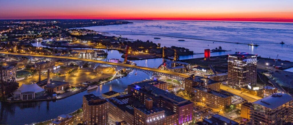 Aerial view of a cityscape at dusk with a river winding through. Bridges, illuminated buildings, and a waterfront area are visible. The sky displays a vibrant sunset with deep orange and purple hues, reflecting on the water.