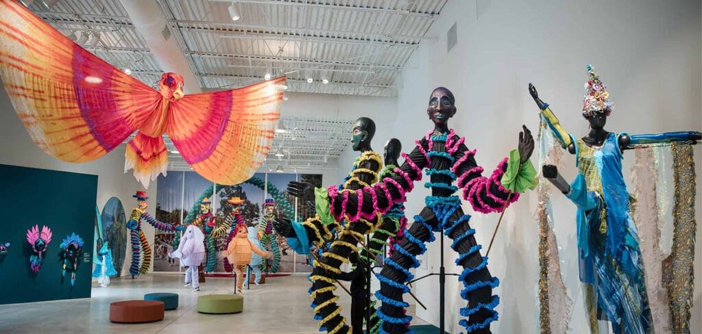 A vibrant art exhibition featuring colorful, whimsical sculptures and large, butterfly-like figures suspended from the ceiling. The space is filled with mannequins wearing elaborate, bright costumes in a spacious, well-lit gallery.