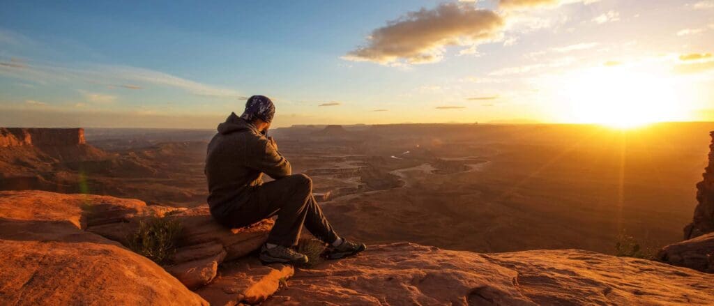 A person sits on a rock, overlooking a vast desert landscape at sunset. The sky is partly cloudy, and the setting sun casts a warm glow over the scene. The person is wearing a hoodie and a bandana, gazing towards the horizon.