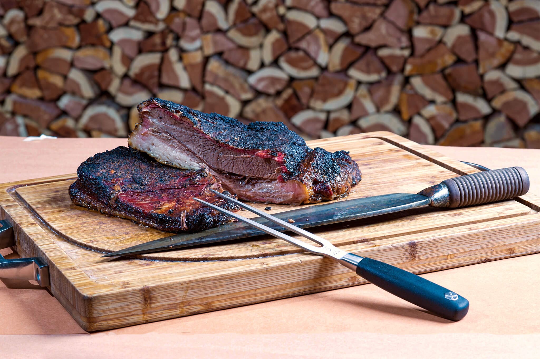 Stack of sliced smoked brisket on a wooden cutting board with a large knife and fork. Background shows stacked firewood.