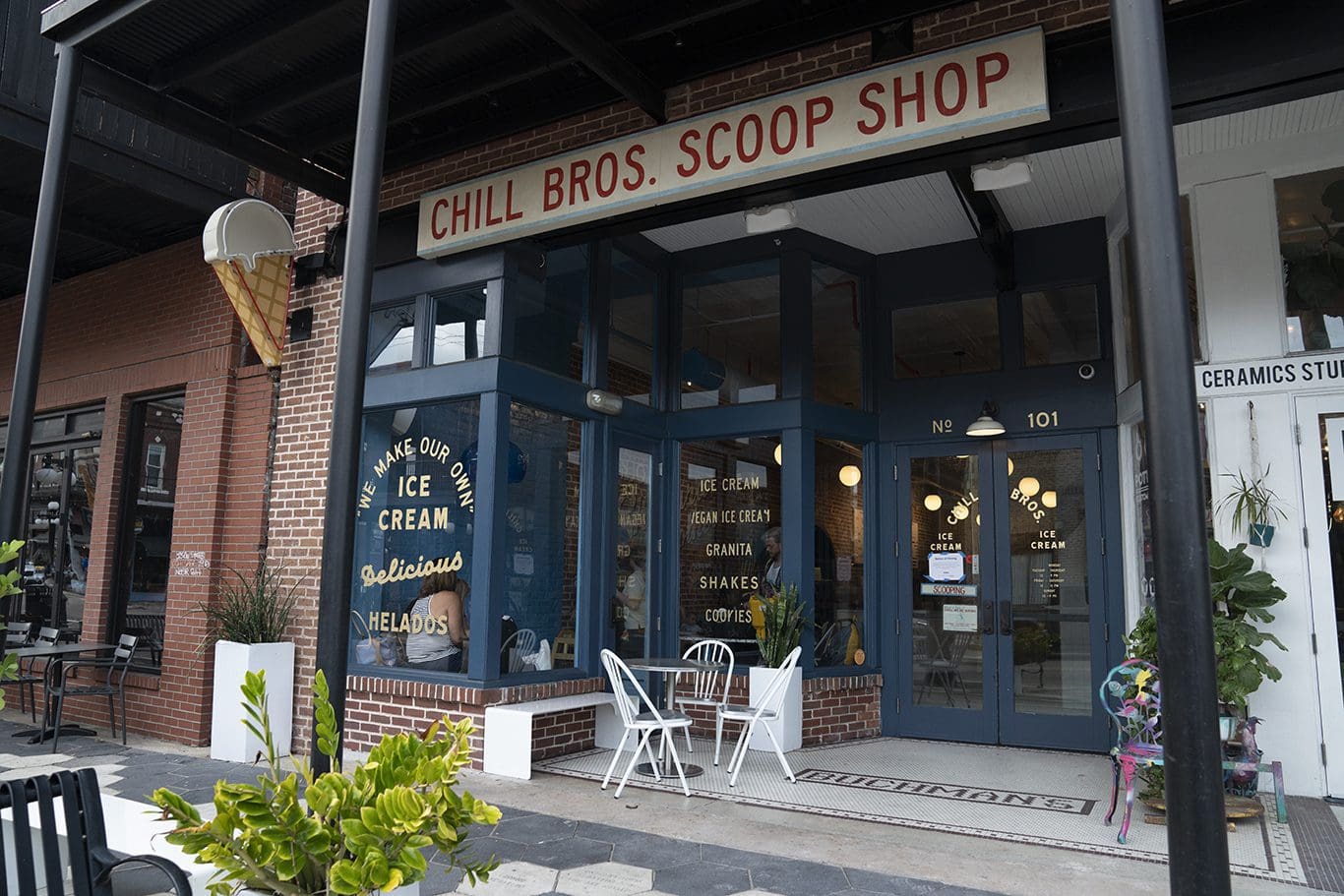 Street view of an ice cream shop named Chill Bros. Scoop Shop. The entrance has large glass windows and doors, with seating outside. A ceramics studio is visible next door. Plants and a decorative sculpture are placed near the entrance.
