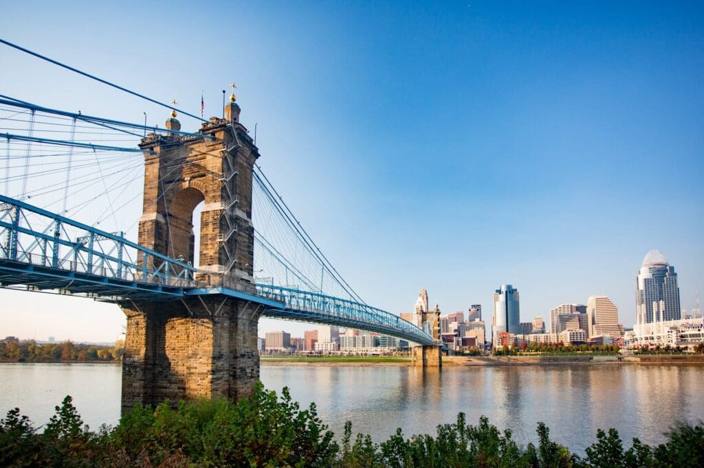 The image shows the John A. Roebling Suspension Bridge spanning across a calm river, connecting to the skyline of Cincinnati. The clear blue sky enhances the view of tall buildings in the background. Lush greenery lines the riverbank.