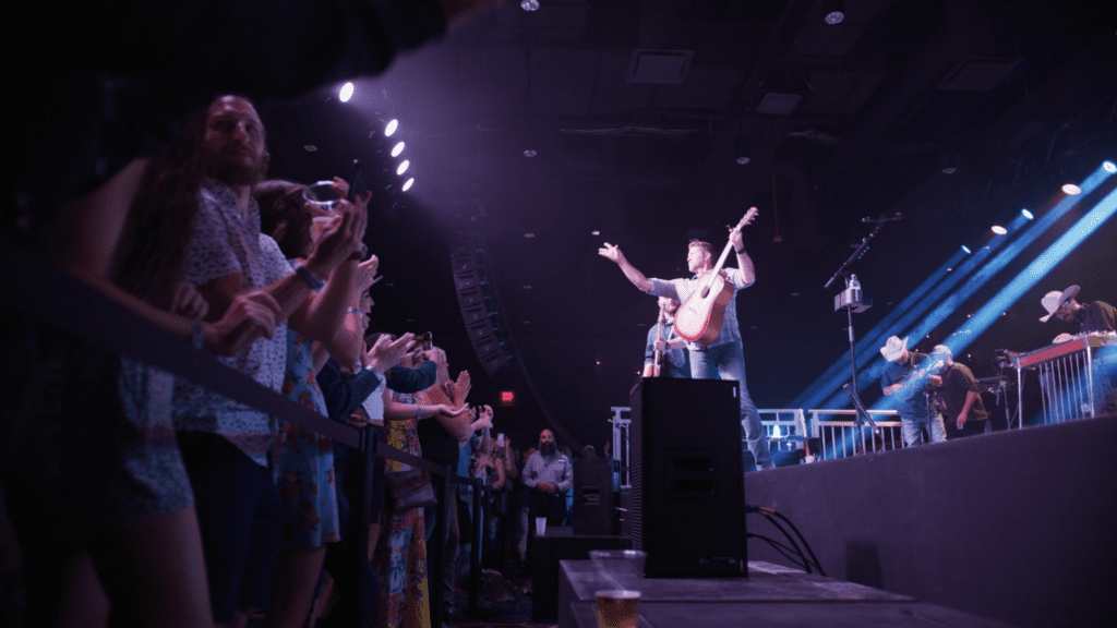 A musician on stage holds a guitar up triumphantly, engaging with a cheering crowd at a lively indoor concert. Bright stage lights illuminate the scene, and fans eagerly capture the moment with their phones.
