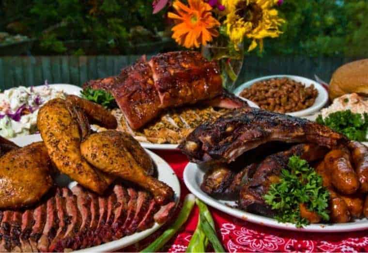 A table laden with barbecued meats including ribs, chicken, and sliced brisket. Sides of coleslaw, baked beans, and a bun accompany the meal. Bright flowers enhance the vibrant presentation. A red bandana cloth serves as the table covering.