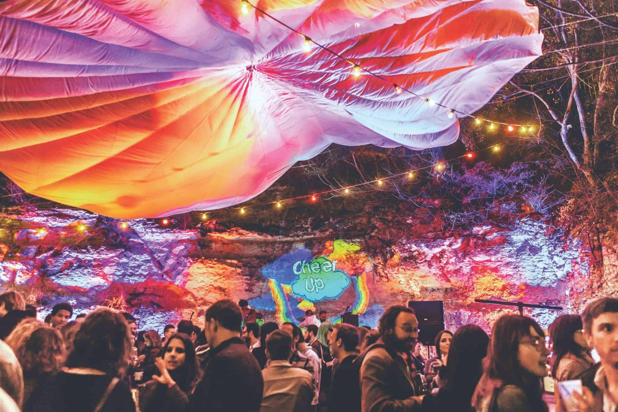 A vibrant outdoor party scene with colorful lights and a large orange and white canopy overhead. People are mingling and chatting. The background shows a rock wall with a bright Cheer Up mural illuminated in multiple colors.