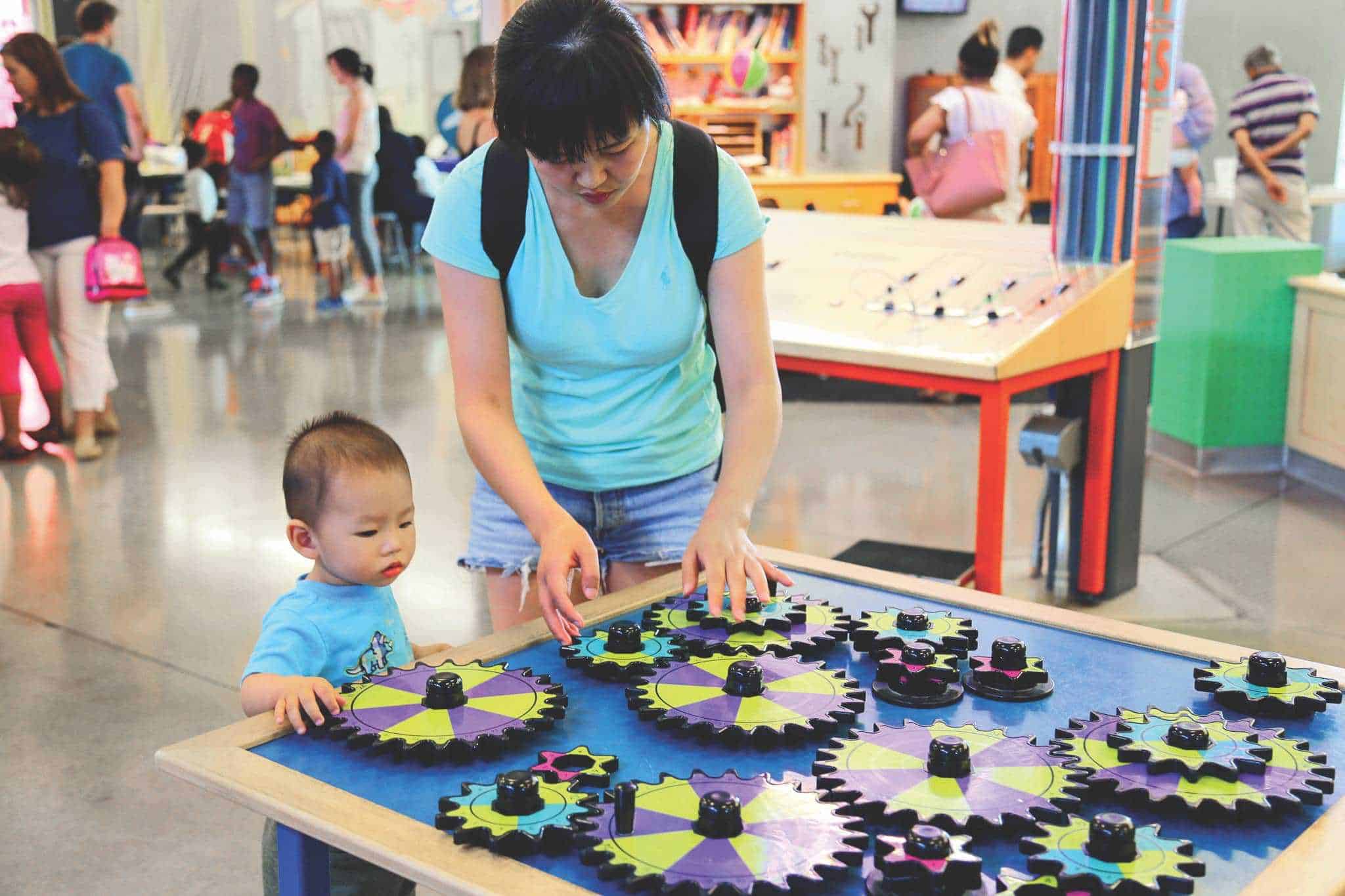 A woman and a young child engage with colorful, interlocking gears on a table at a museum or interactive play area. The room is bustling with other visitors, and various exhibits are visible in the background.