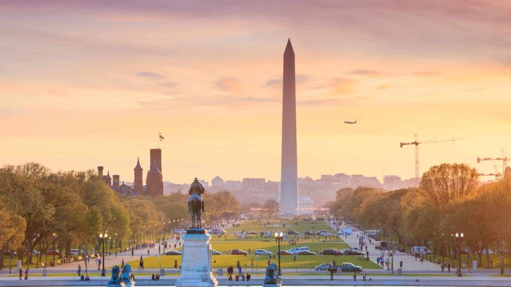 View of the Washington Monument at sunset, with green lawns and pathways lined by trees. A statue on horseback stands in the foreground. Cars and people are visible, with a plane flying in the sky and cranes in the distance.