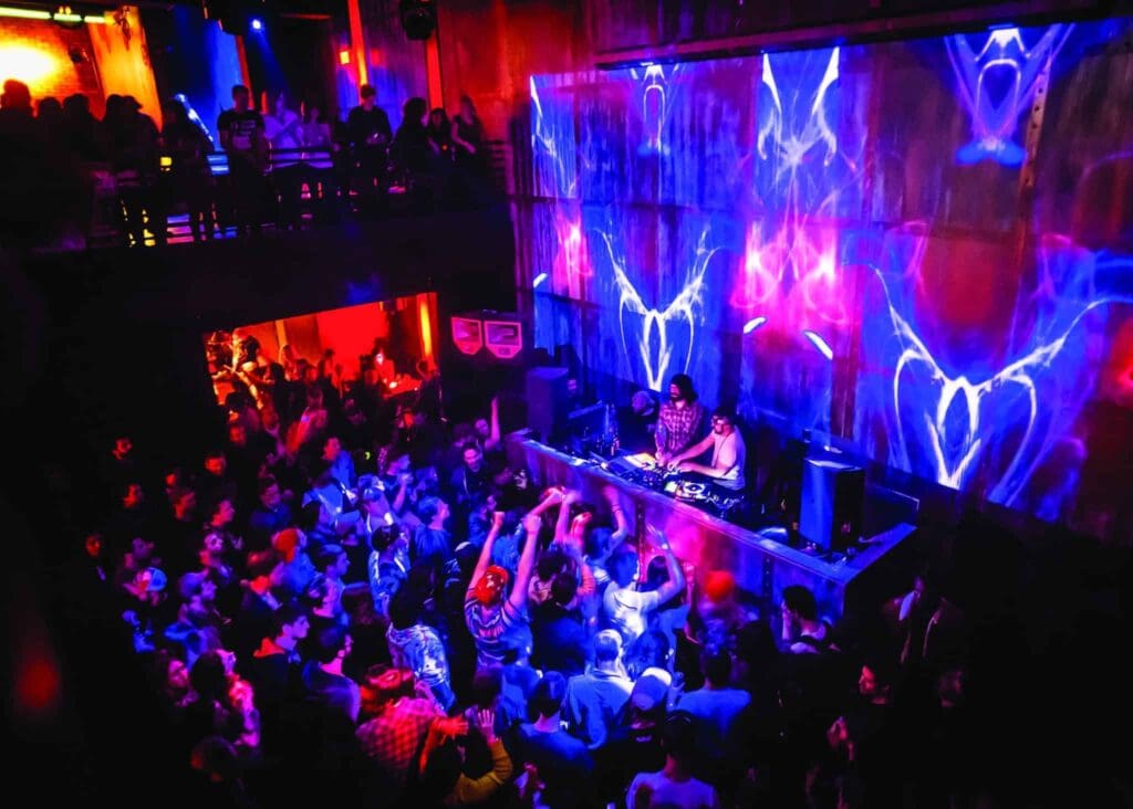 A vibrant nightclub scene with a DJ performing on stage. Colorful lights and visuals illuminate the wall, casting vivid blue and purple patterns. A packed crowd is dancing energetically on the floor, creating a lively and dynamic atmosphere.