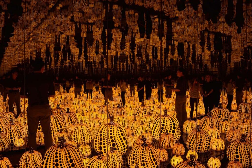 A room filled with glowing pumpkin sculptures covered in black dots. The walls and ceiling are mirrored, creating an infinite reflection of the pumpkins. People stand among the pumpkins, adding to the immersive experience.