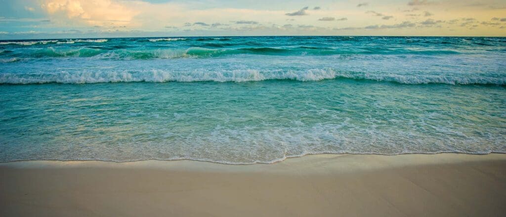 A serene beach scene with gentle waves lapping at the sandy shore. The sea is a vibrant shade of turquoise under a partly cloudy sky, with the horizon blending seamlessly into the distance. The light reflects softly on the waters surface.