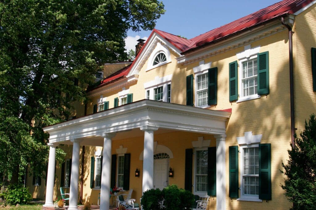 A large yellow house with a red roof, white columns, and a grand front porch. The windows have white shutters, and there are trees providing shade around the house. Two chairs and small plants are on the porch.