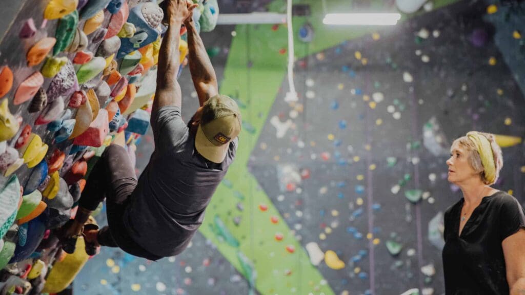 A person wearing a cap climbs an indoor rock wall with colorful holds, while another person stands nearby watching. The background features more climbing routes and bright lighting.