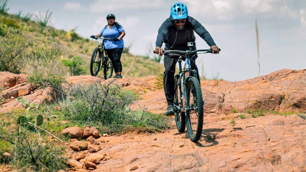 Two people are mountain biking on a rocky trail. The person in the foreground wears a blue helmet and black attire, while the person behind wears a green helmet and blue shirt. Vegetation surrounds the trail under a cloudy sky.