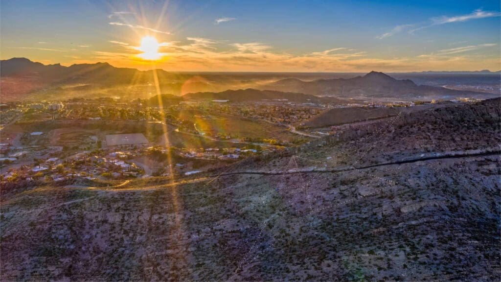 Sunset over a mountainous landscape with a city nestled in the valley. Sun rays create a warm glow across the scene, highlighting the contours of the hills. A mix of nature and urban landscape under a clear evening sky.