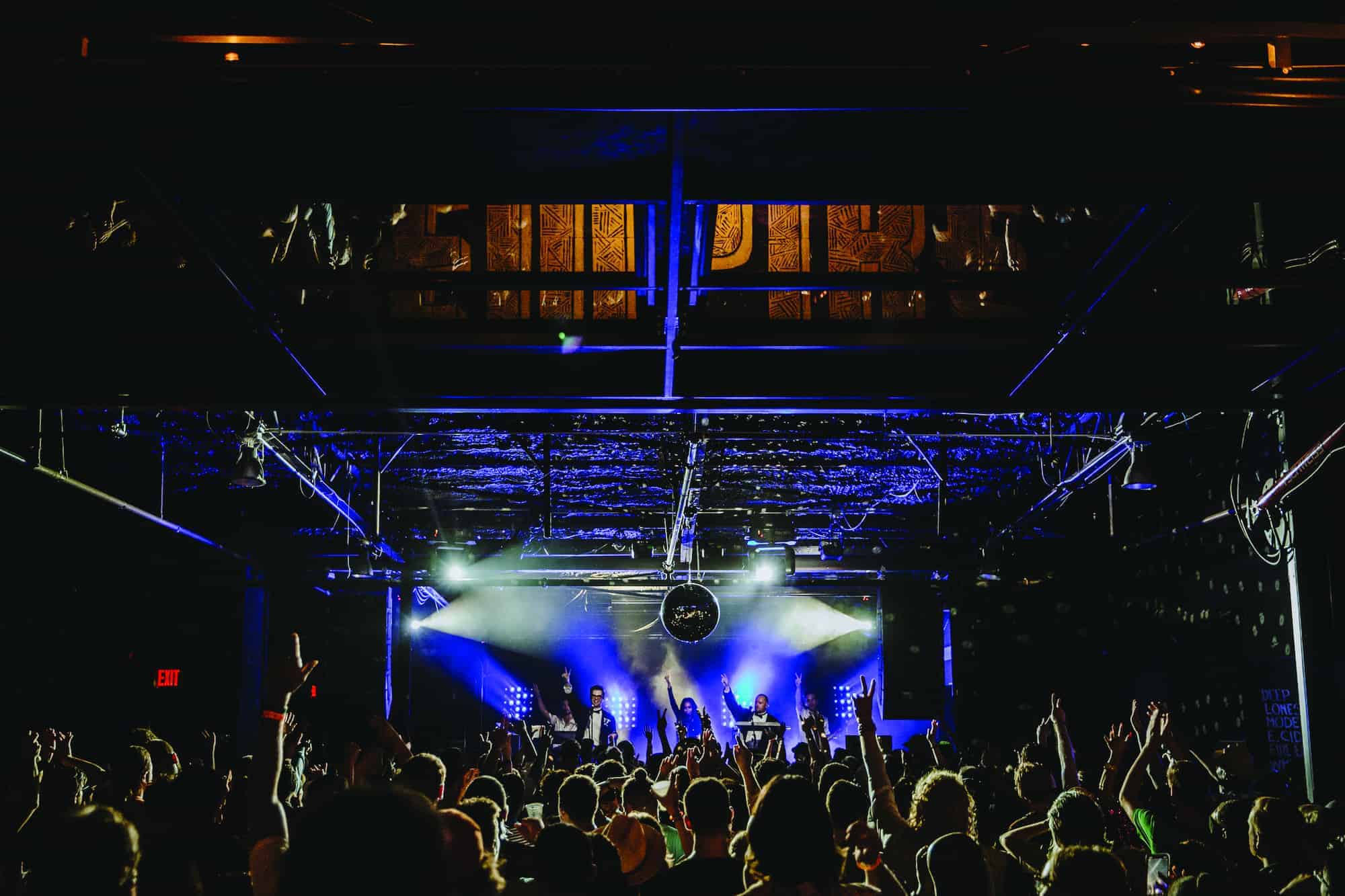 A vibrant concert scene in a dimly lit venue, with a band performing on stage. Colorful lights illuminate the band, casting a glow over the enthusiastic crowd. Hands are raised in the air, creating an energetic atmosphere.