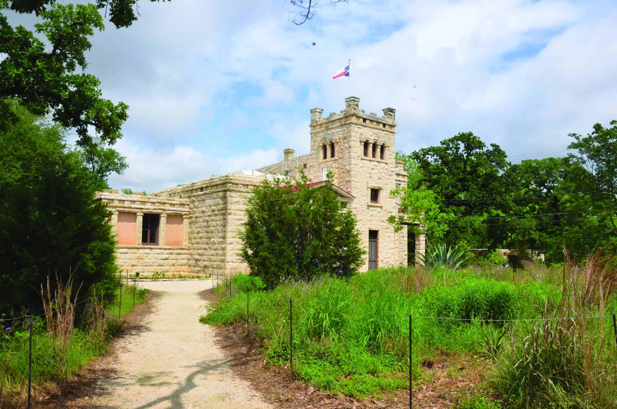 A stone castle-like building with a prominent Texas flag on the roof, surrounded by lush greenery and a dirt path leading to it. The sky is partly cloudy.