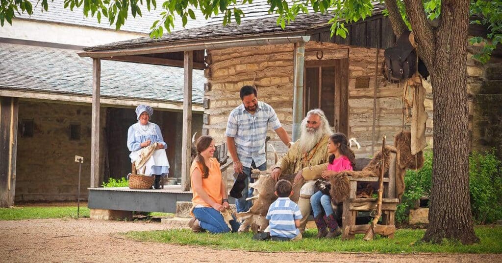 A group of people, including an older man with a long beard, is gathered in front of a rustic log cabin. They are examining animal pelts. A woman in period clothing stands nearby, holding a basket. The setting appears historical.