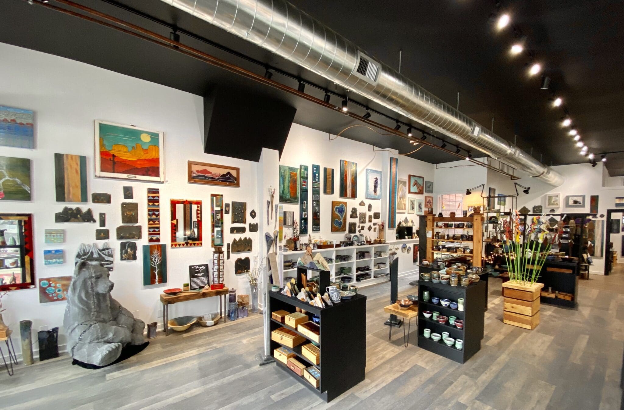 A spacious art gallery with wooden flooring and a dark ceiling featuring ductwork. The walls display a variety of paintings and artworks. Shelves and tables showcase pottery and decorative items. A large sculpture is visible on the left.