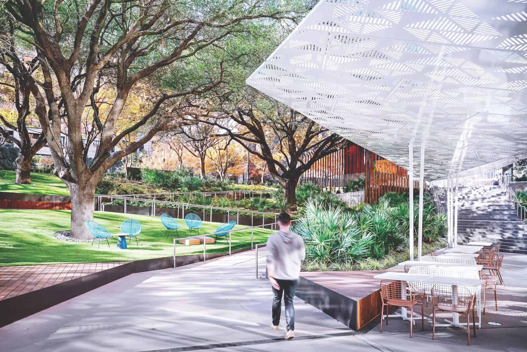 A person walks along a path in a sunlit park. The area features modern outdoor seating, large trees, and landscaped greenery. A geometric-patterned canopy provides shade over some tables and chairs.
