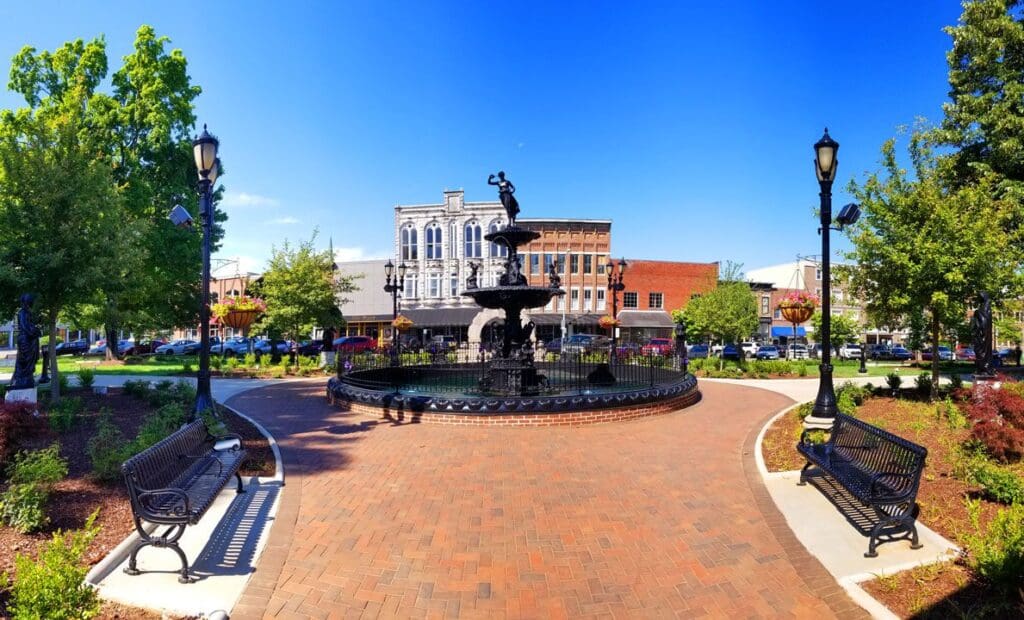 A town square with a central fountain surrounded by a black wrought-iron fence. Brick pathways lead to benches and lamp posts. In the background, there are historic buildings with shops and trees lining the street under a clear blue sky.