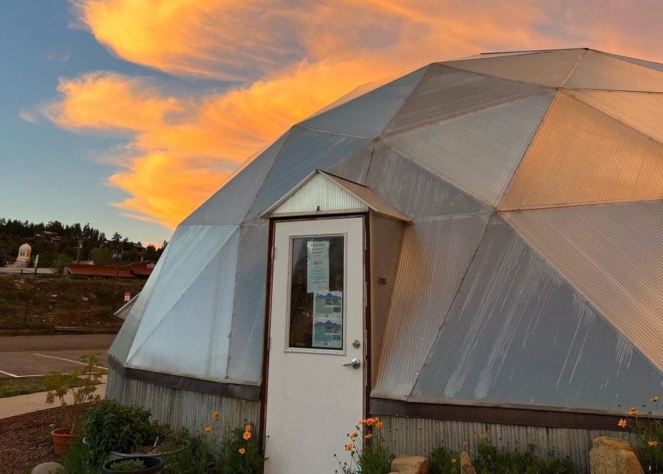 A geodesic dome with a white door set against a vibrant sunset sky. The clouds are lit in orange and gold hues. Surrounding the dome are small plants and a parking area.