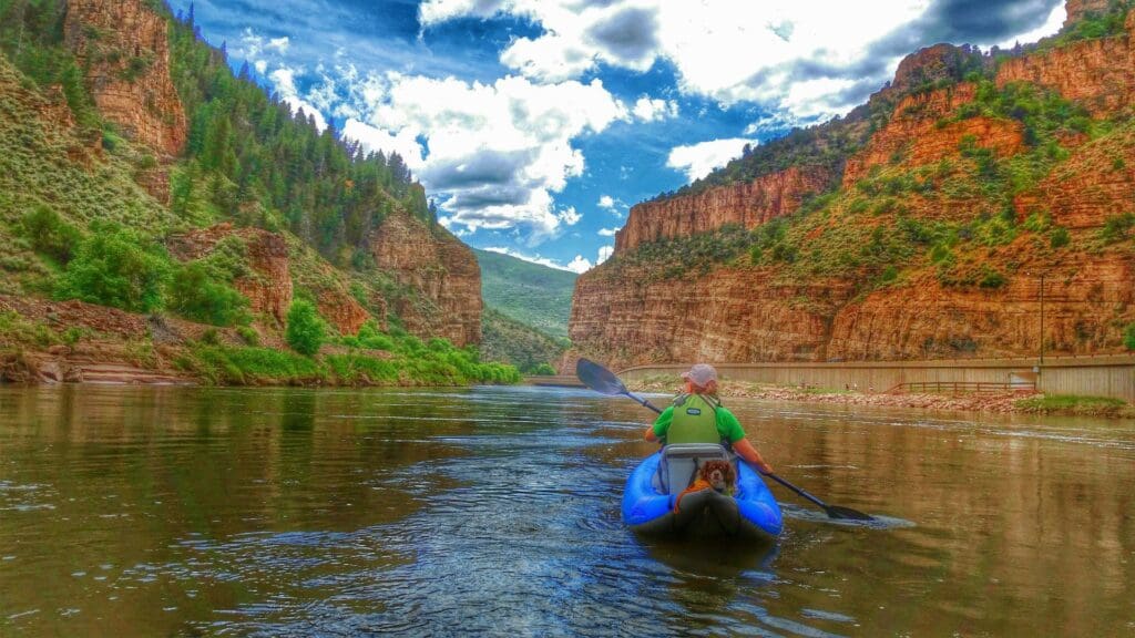 A person in a blue kayak paddles down a calm river between striking red rock cliffs and green foliage under a partly cloudy sky. The scene is tranquil, showcasing the grandeur of the natural canyon landscape.