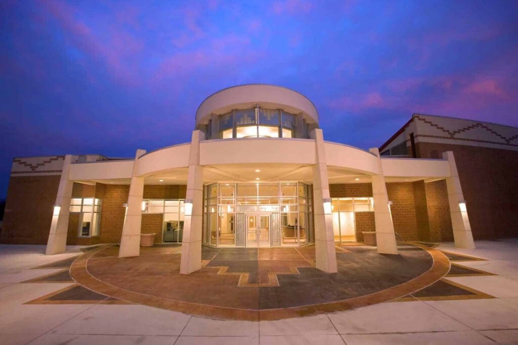 A modern building with a circular entrance is illuminated against a twilight sky. The structure features large glass windows and geometric architectural details. The ground has a pattern of concentric circles leading to the entrance.