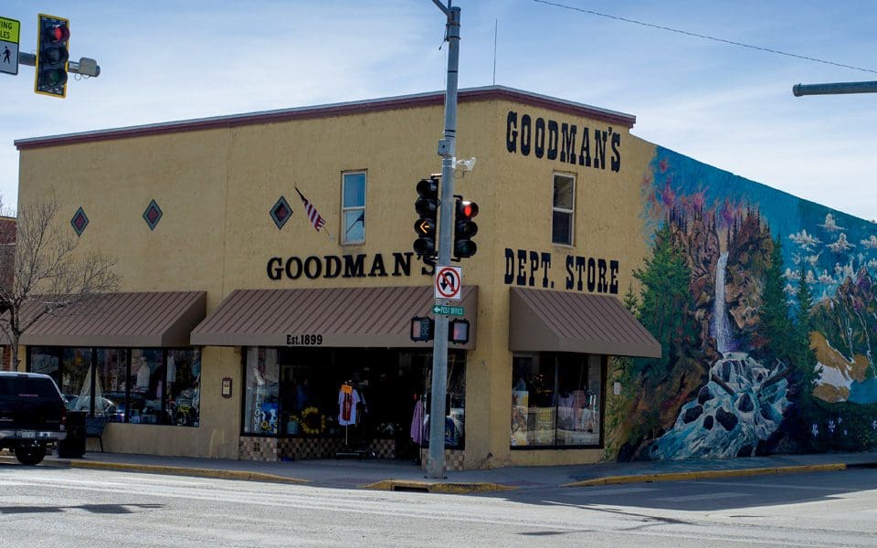 A beige building with brown awnings houses Goodmans Dept. Store, established in 1899. A colorful mural of trees and rocks adorns the side wall. Traffic lights and a no-left-turn sign are visible at the corner.